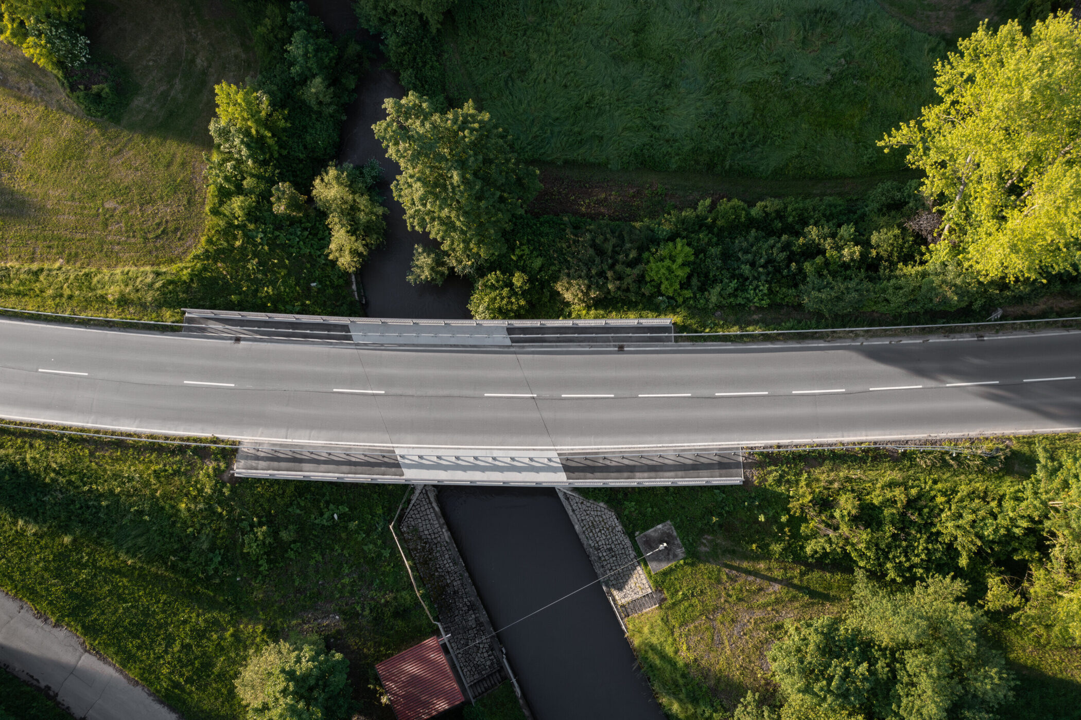 Straße überquert Fluss, umgeben von Bäumen und Wiesen, aus der Vogelperspektive.