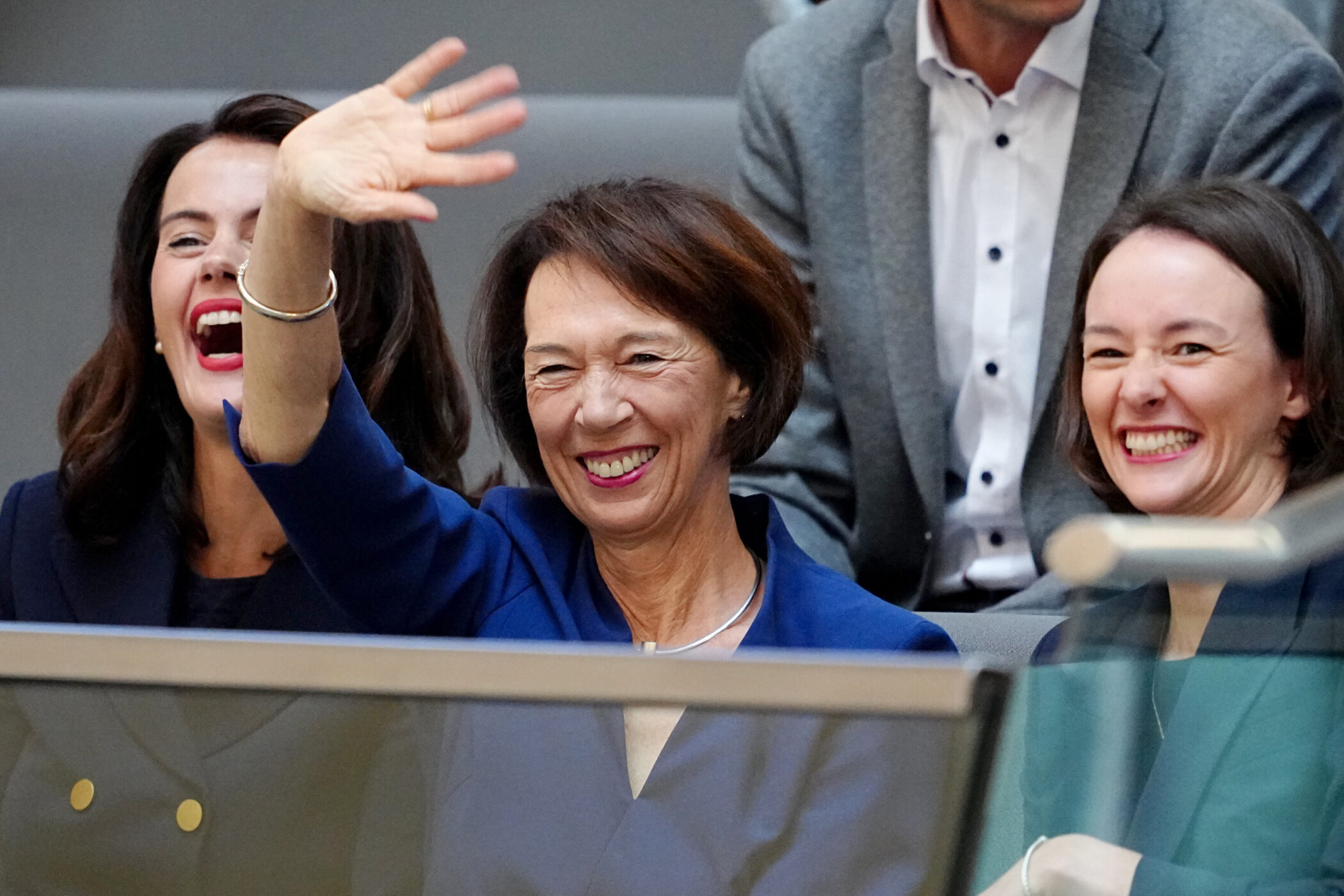 Drei lachende Frauen, eine winkt, im Hintergrund ein Mann in grauem Sakko.