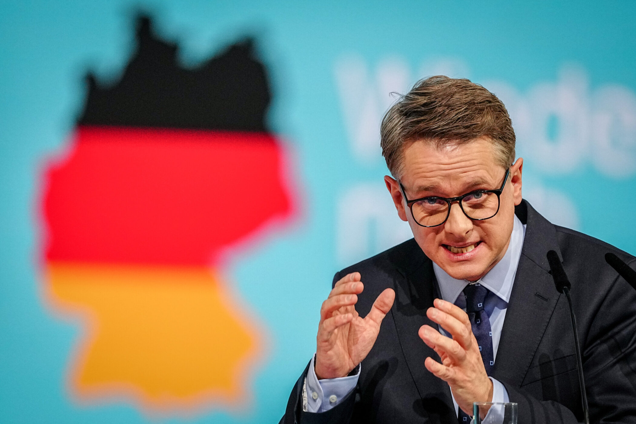 Mann mit Brille spricht, Deutschlandkarte im Hintergrund.