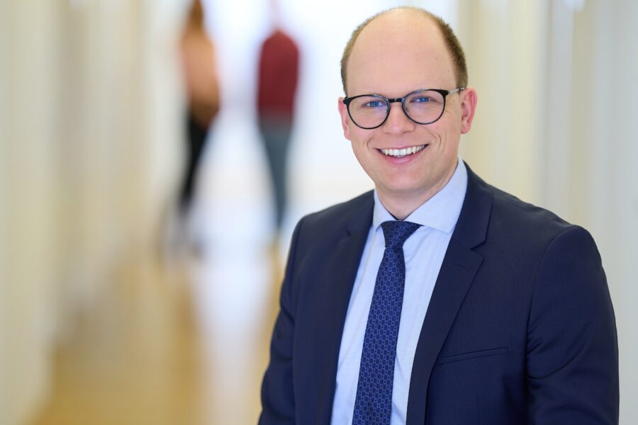 Lächelnder Mann mit Brille und Anzug in einem Flur, zwei unscharfe Personen im Hintergrund.