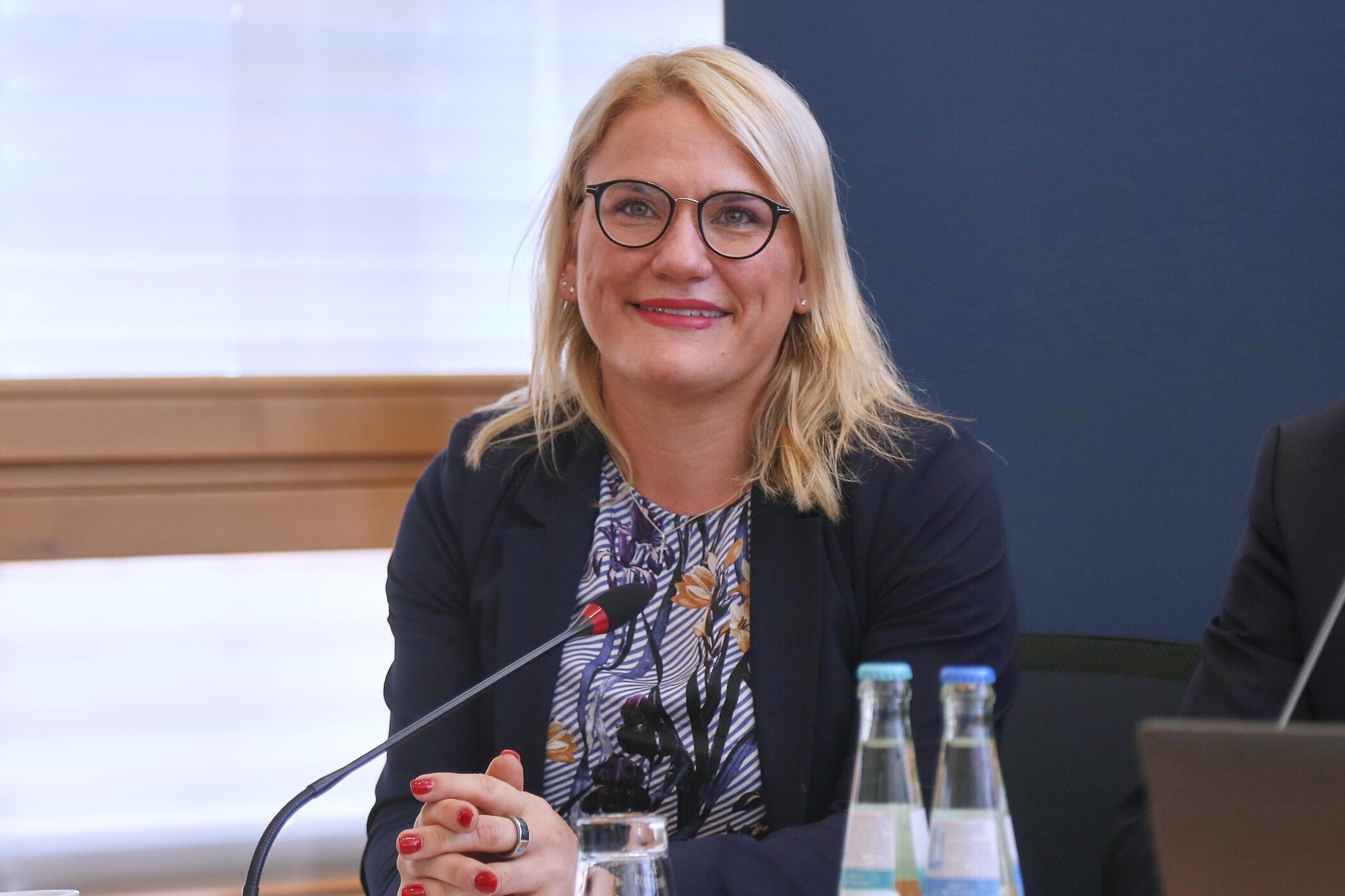 Eine Frau mit Brille sitzt lächelnd an einem Tisch mit Mikrofon.