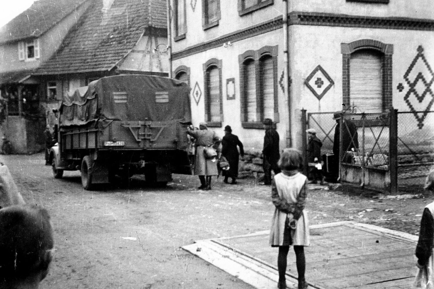 Lkw mit Plane, mehrere Personen auf Straße, Gebäude im Hintergrund, Kind im Vordergrund.