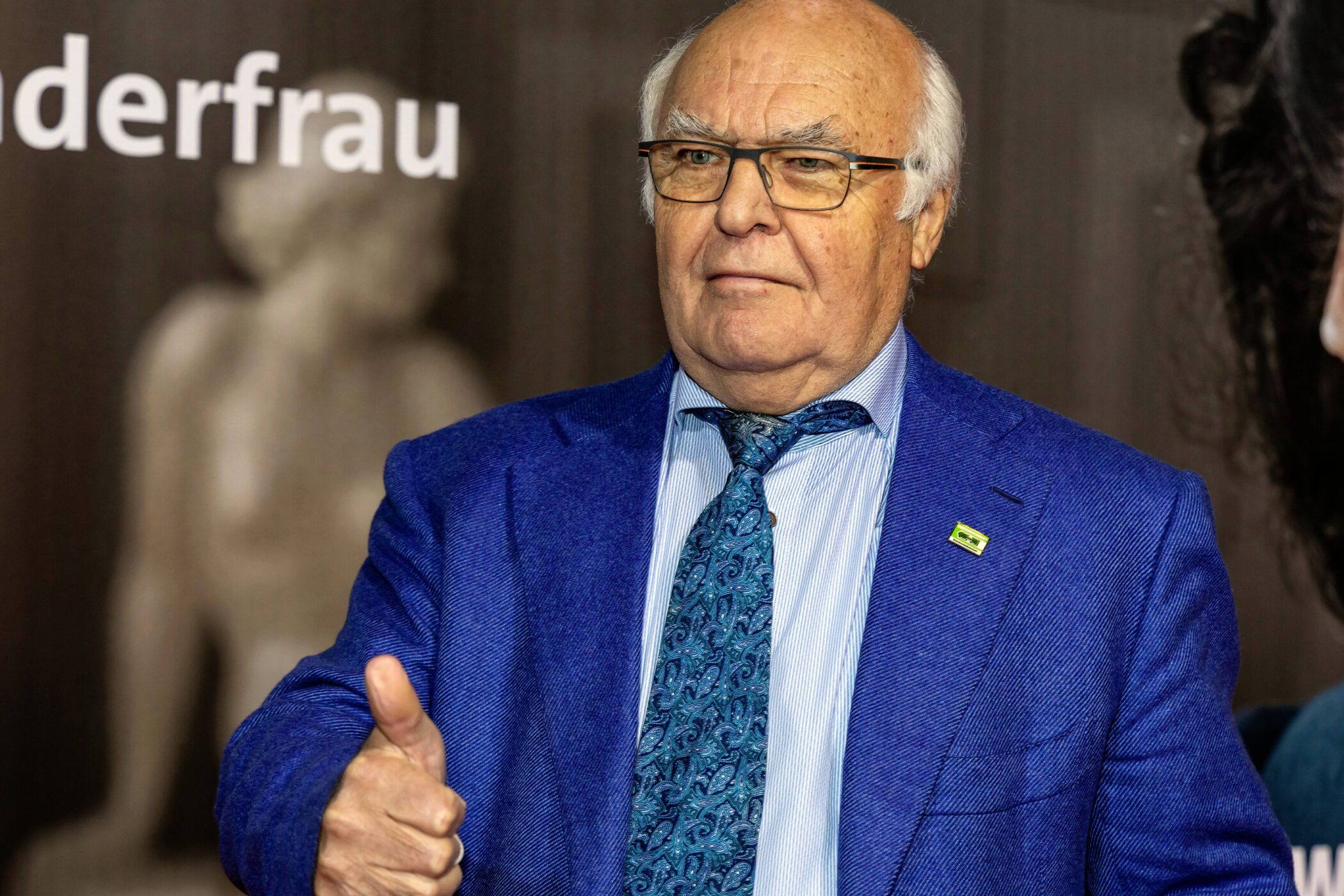 Älterer Mann im blauen Anzug zeigt Daumen hoch, unscharfer Hintergrund.