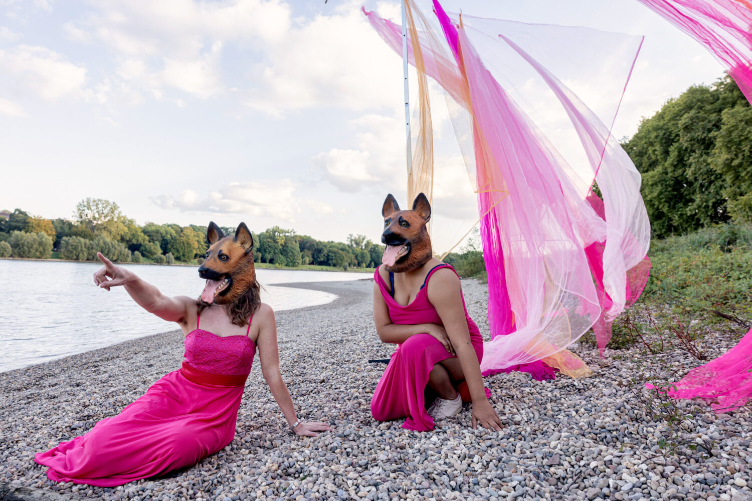 Zwei Personen in pinken Kleidern mit Hundemasken am Kiesstrand, rosa Stoffe im Hintergrund.