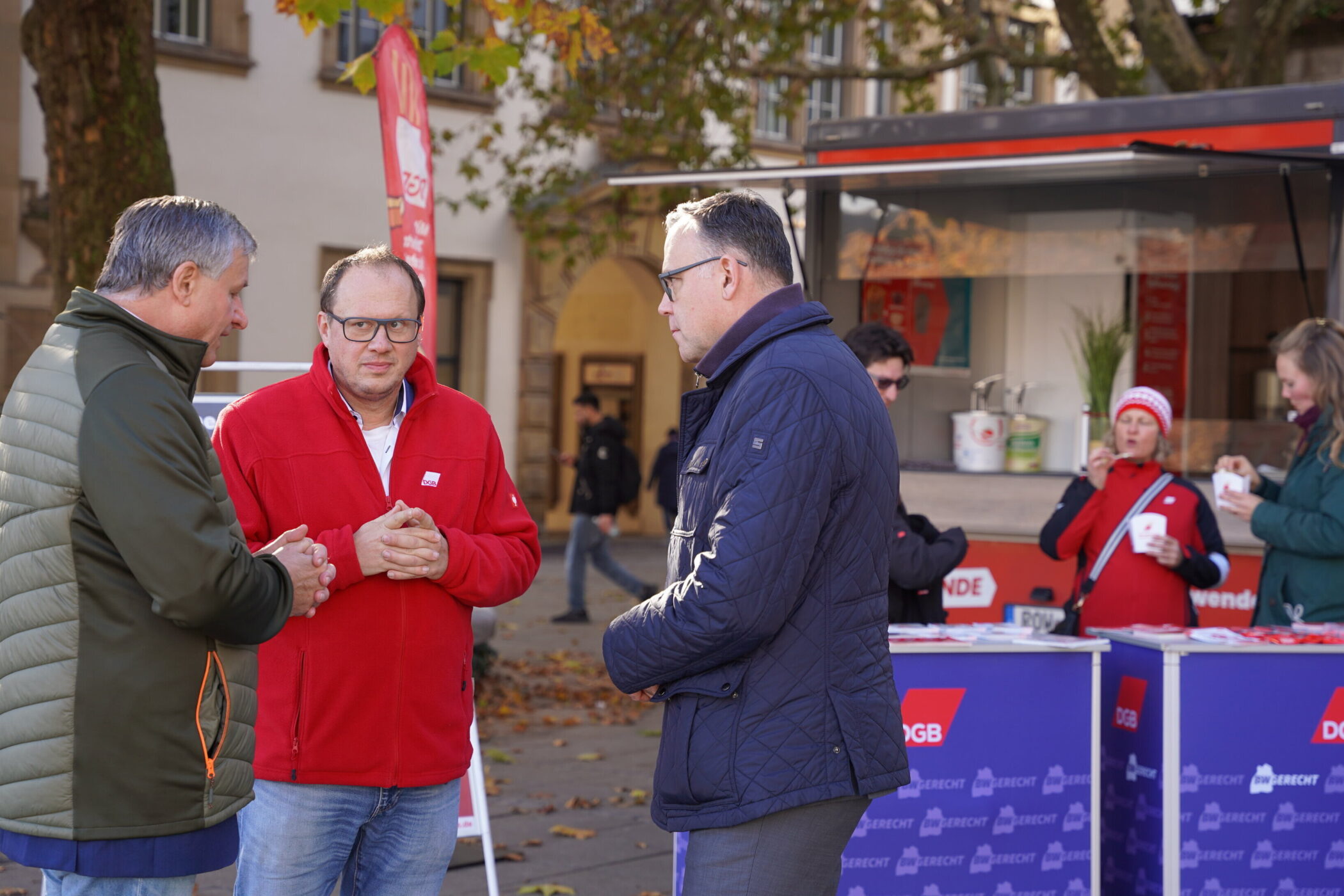 Drei Männer im Gespräch, zwei Frauen essen vor einem Infostand im Freien.