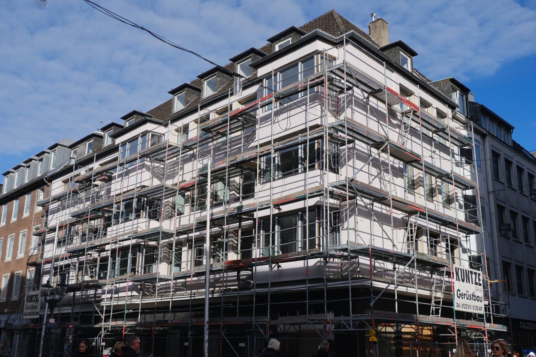 Mehrstöckiges Gebäude mit Baugerüst, Menschen gehen vorbei.