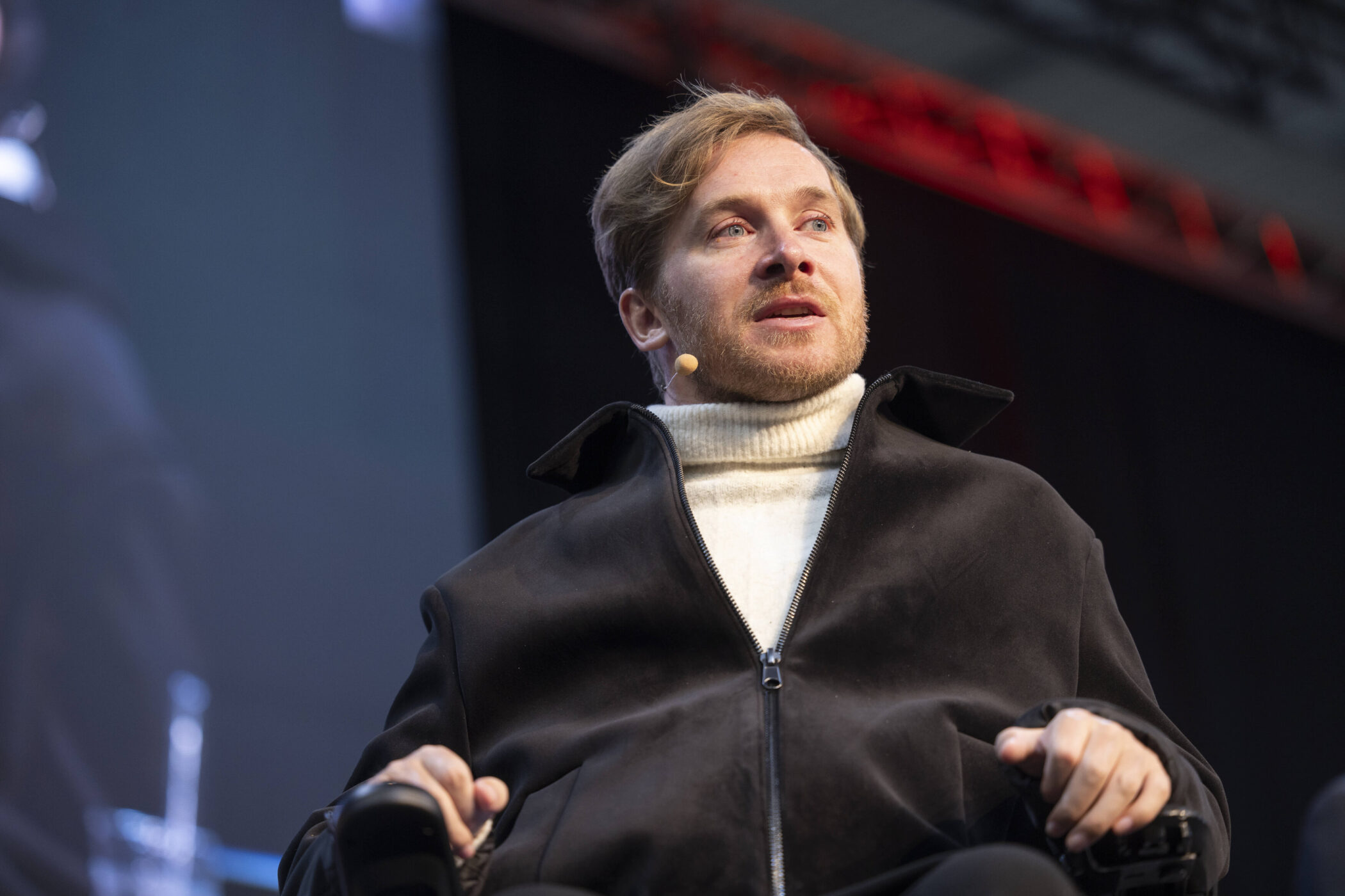 Mann mit Mikrofon am Ohr, trägt dunkle Jacke und Rollkragenpullover.