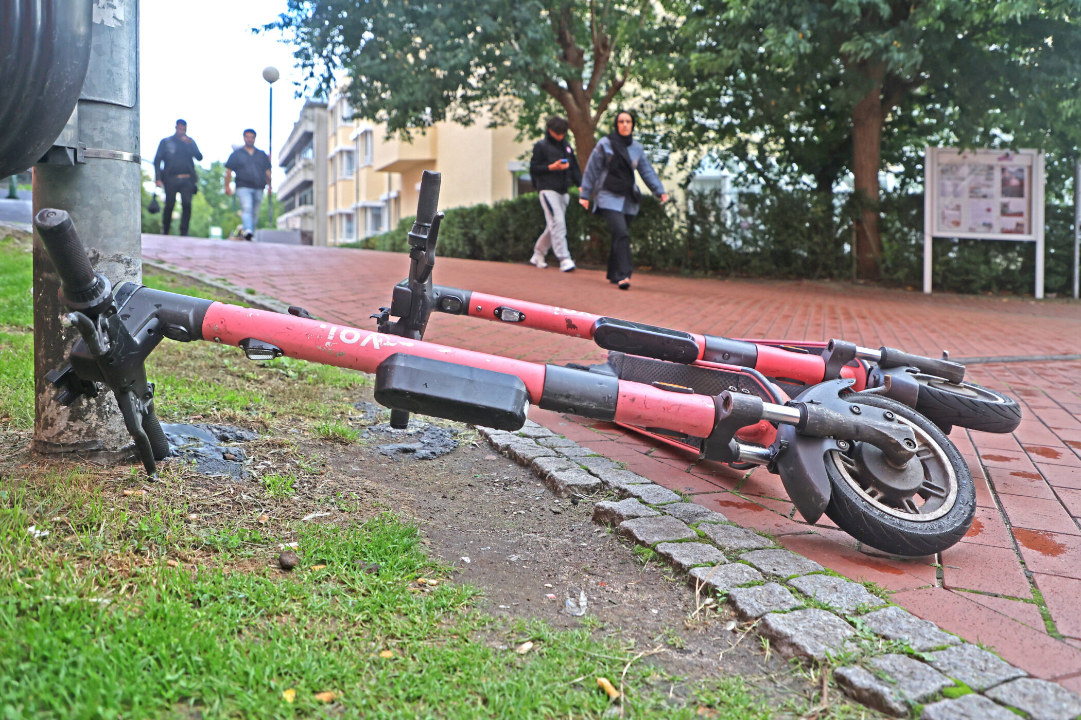 Zwei umgekippte E-Scooter auf Gehweg, Personen im Hintergrund.