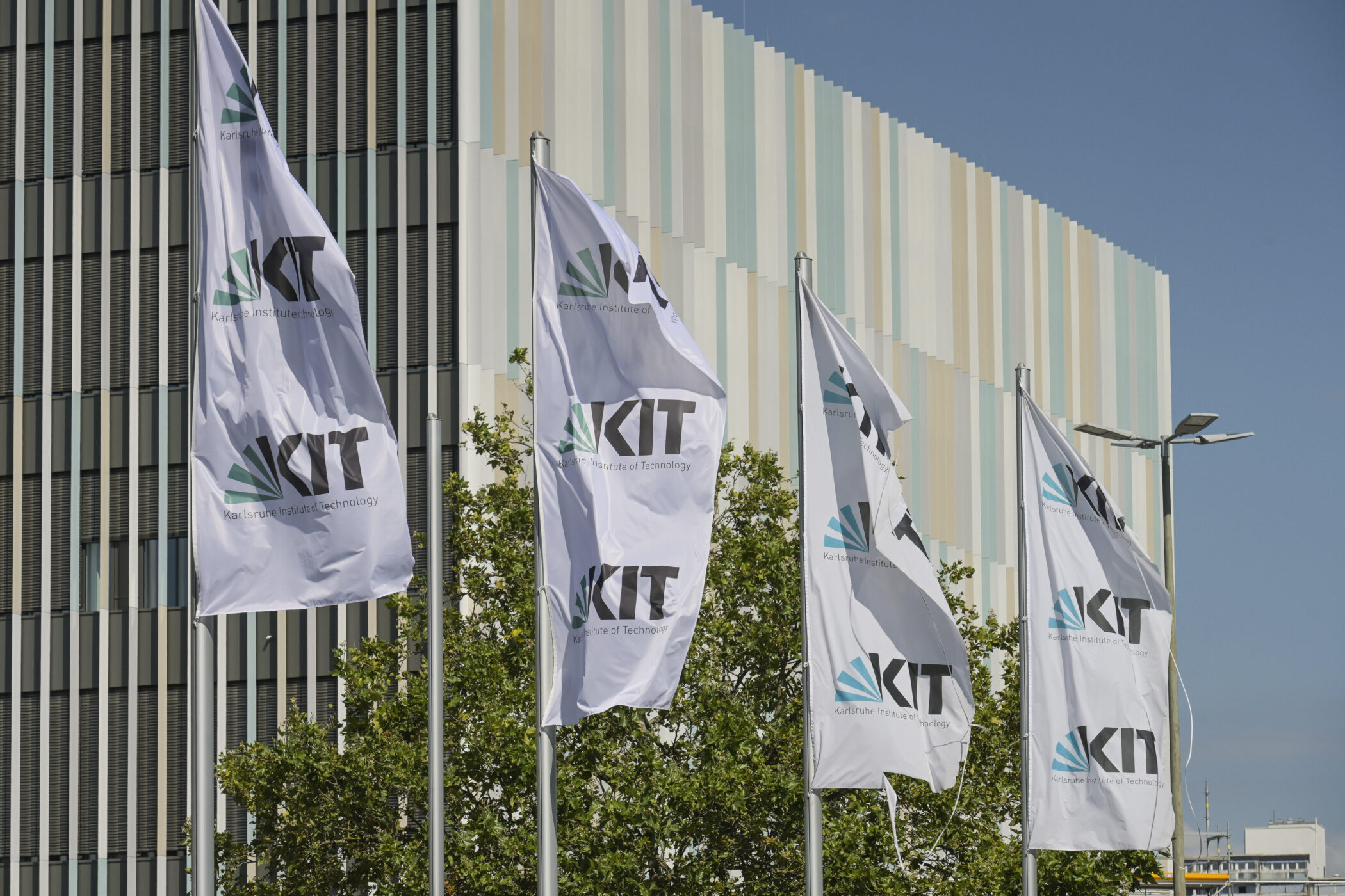 Sechs weiße Fahnen mit KIT-Logo vor modernem Gebäude.