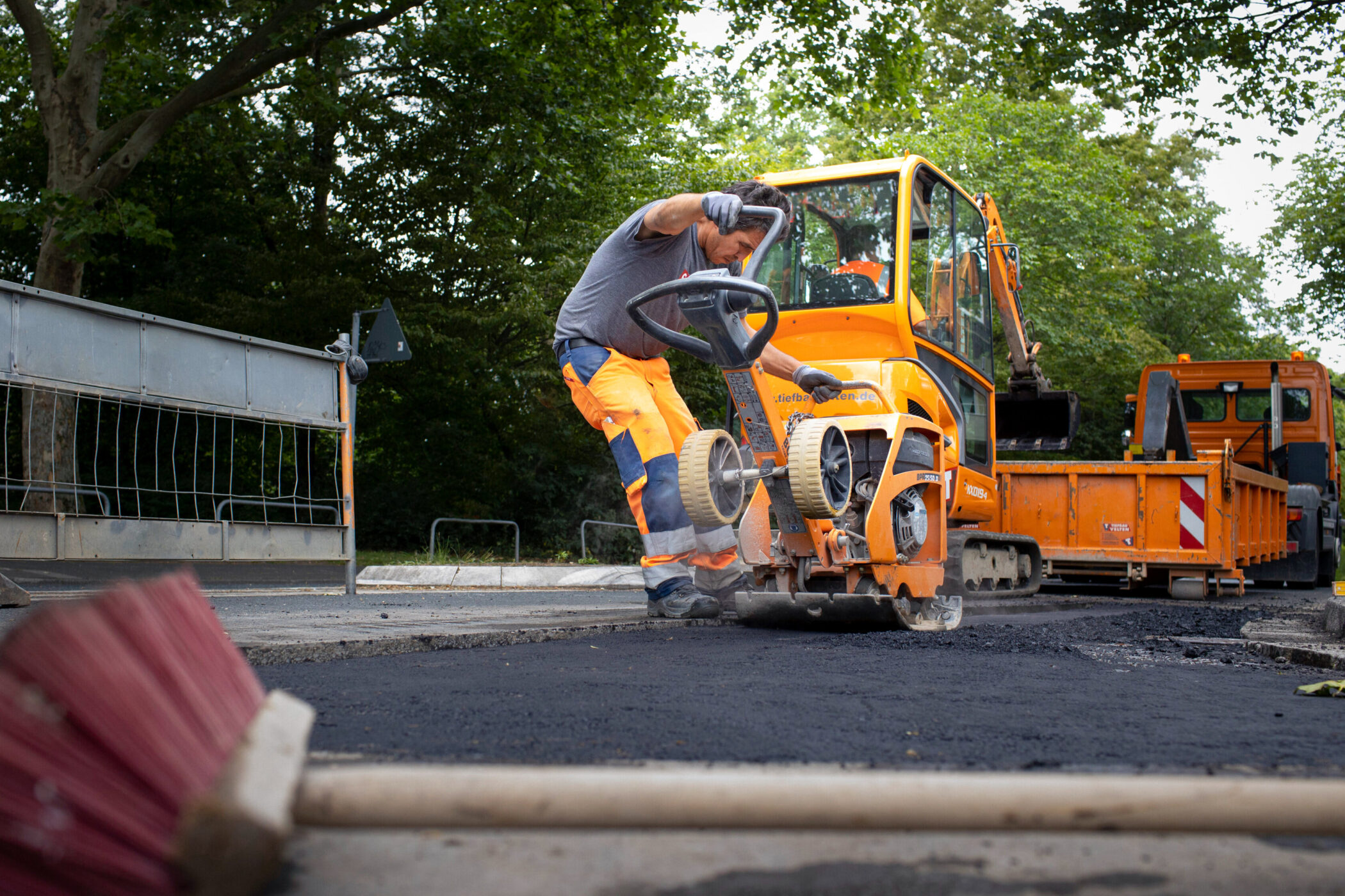 Arbeiter mit Straßenfräse auf Baustelle, orangefarbene Maschinen im Hintergrund.