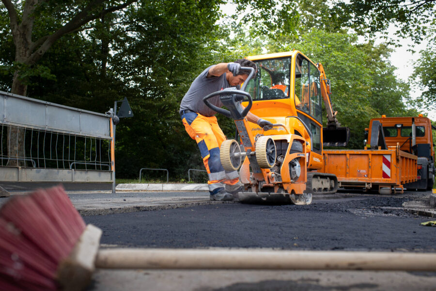 Arbeiter mit Straßenfräse auf Baustelle, orangefarbene Maschinen im Hintergrund.