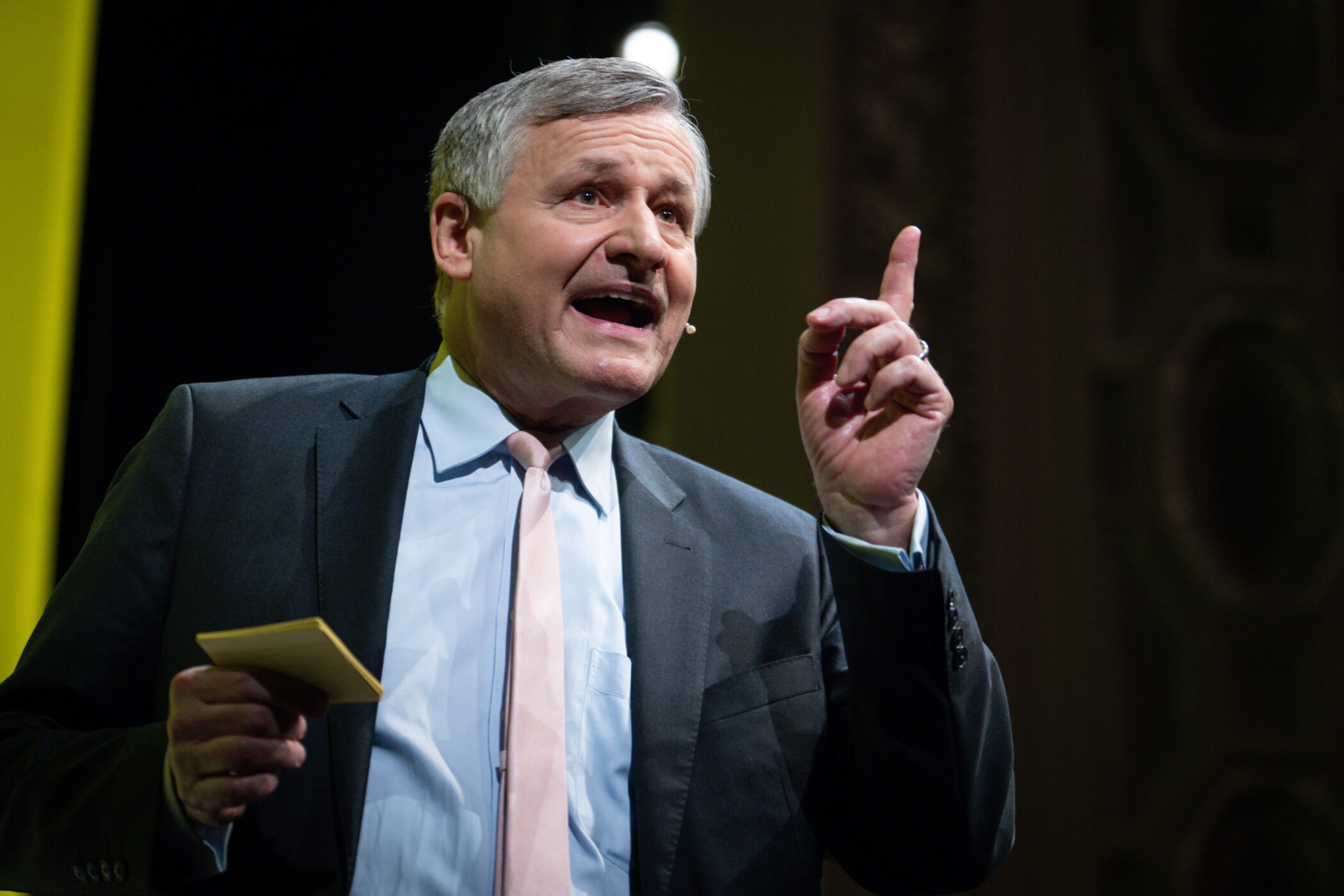 Älterer Mann im Anzug spricht, hebt einen Finger, hält Notizblock.