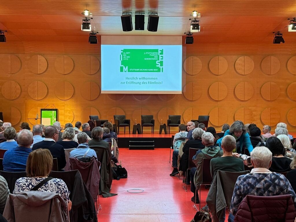 Publikum in einem Saal, Bühne mit Stühlen, Leinwand mit Begrüßungstext.