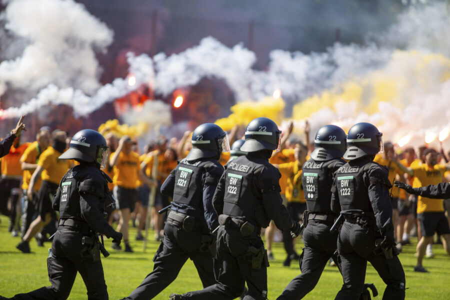 Polizisten in Schutzkleidung laufen, Menschenmenge mit Rauch im Hintergrund.