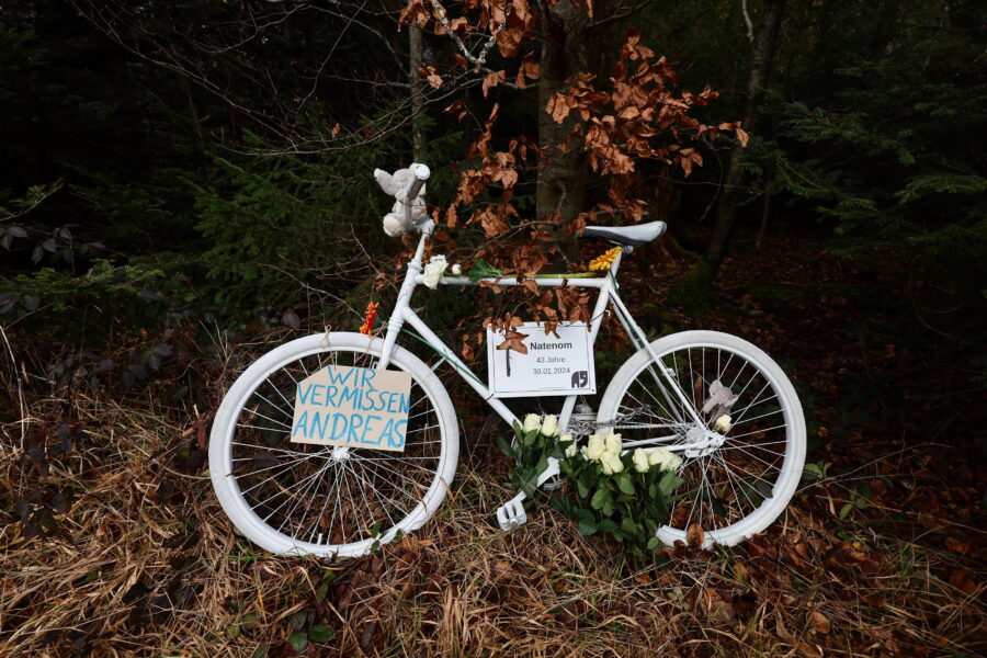 Weißes Fahrrad mit Blumen und Schildern im Wald, Text: "Wir vermissen Andreas".