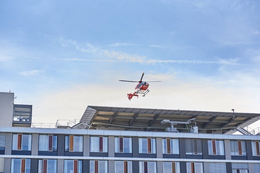 Roter Hubschrauber über einem Gebäude mit Dachplattform, blauer Himmel im Hintergrund.