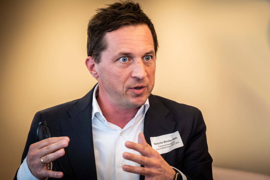 Mann im Anzug spricht, hält Brille in der Hand, Namensschild auf Jacke.