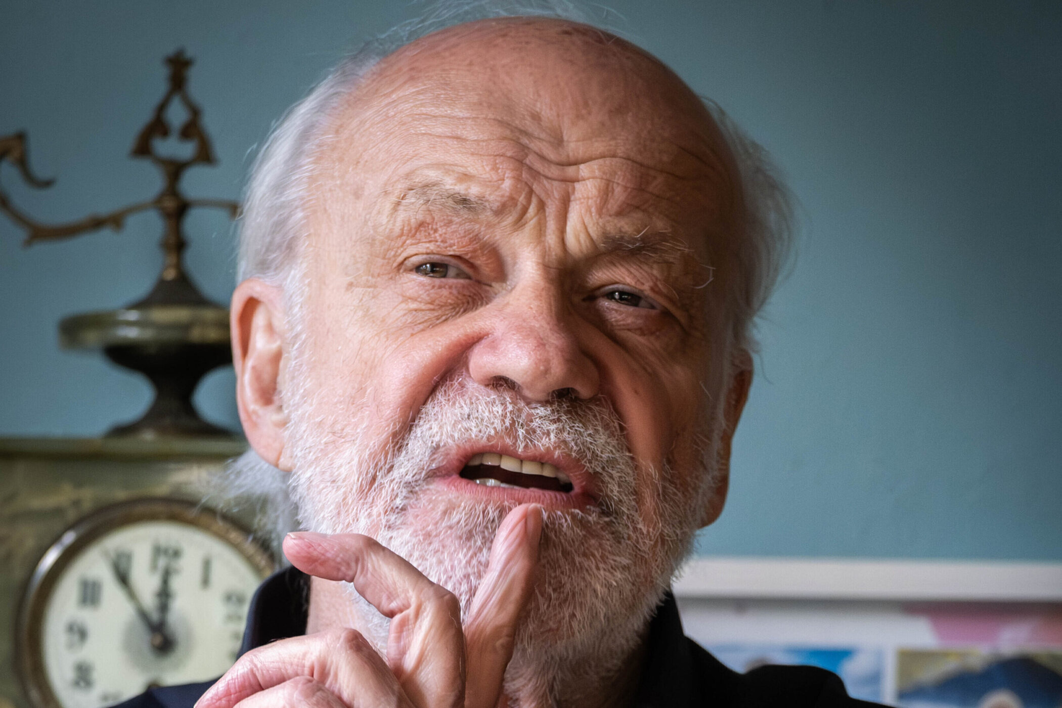Älterer Mann mit Bart, hebt Finger an den Mund, im Hintergrund eine Uhr.
