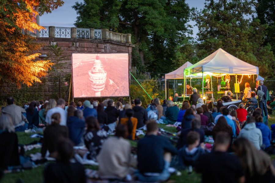 Menschen sitzen im Freien vor einer Leinwand und einem beleuchteten Zelt.