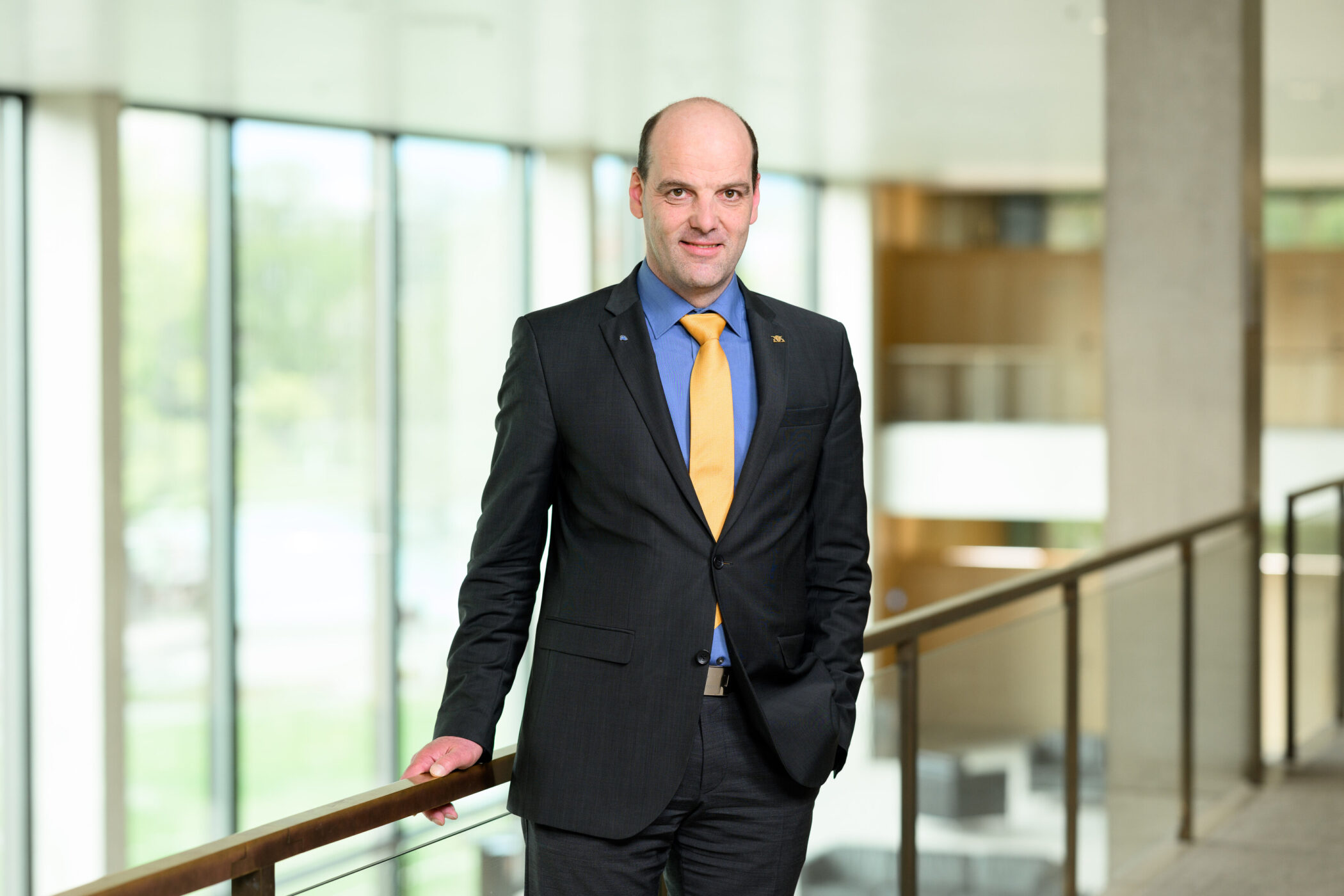Mann im Anzug mit gelber Krawatte steht in modernem Gebäude an einem Geländer.