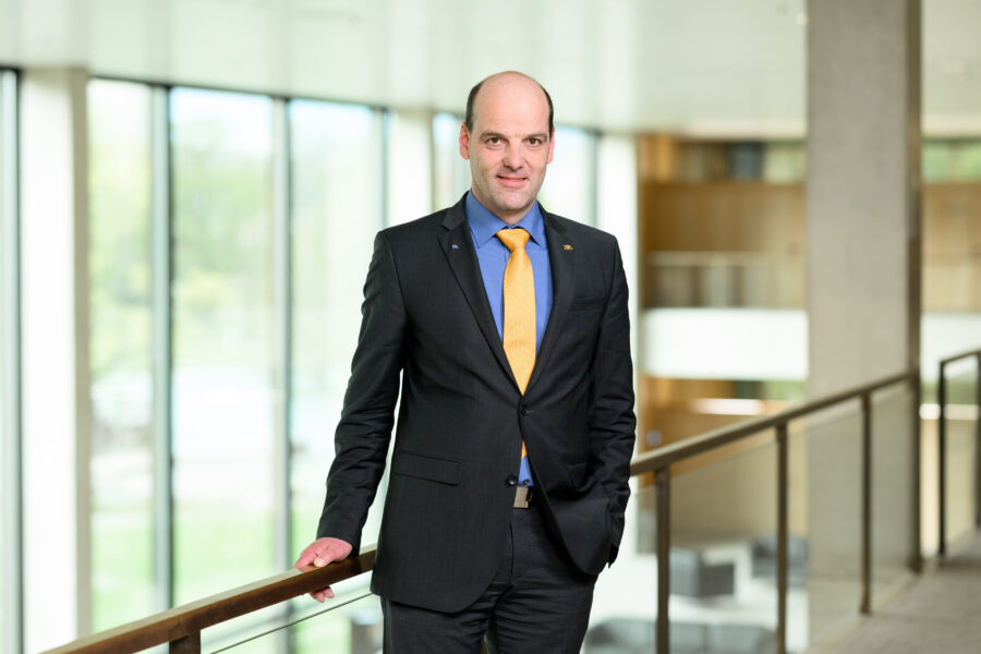 Mann im Anzug mit gelber Krawatte steht in modernem Gebäude an einem Geländer.