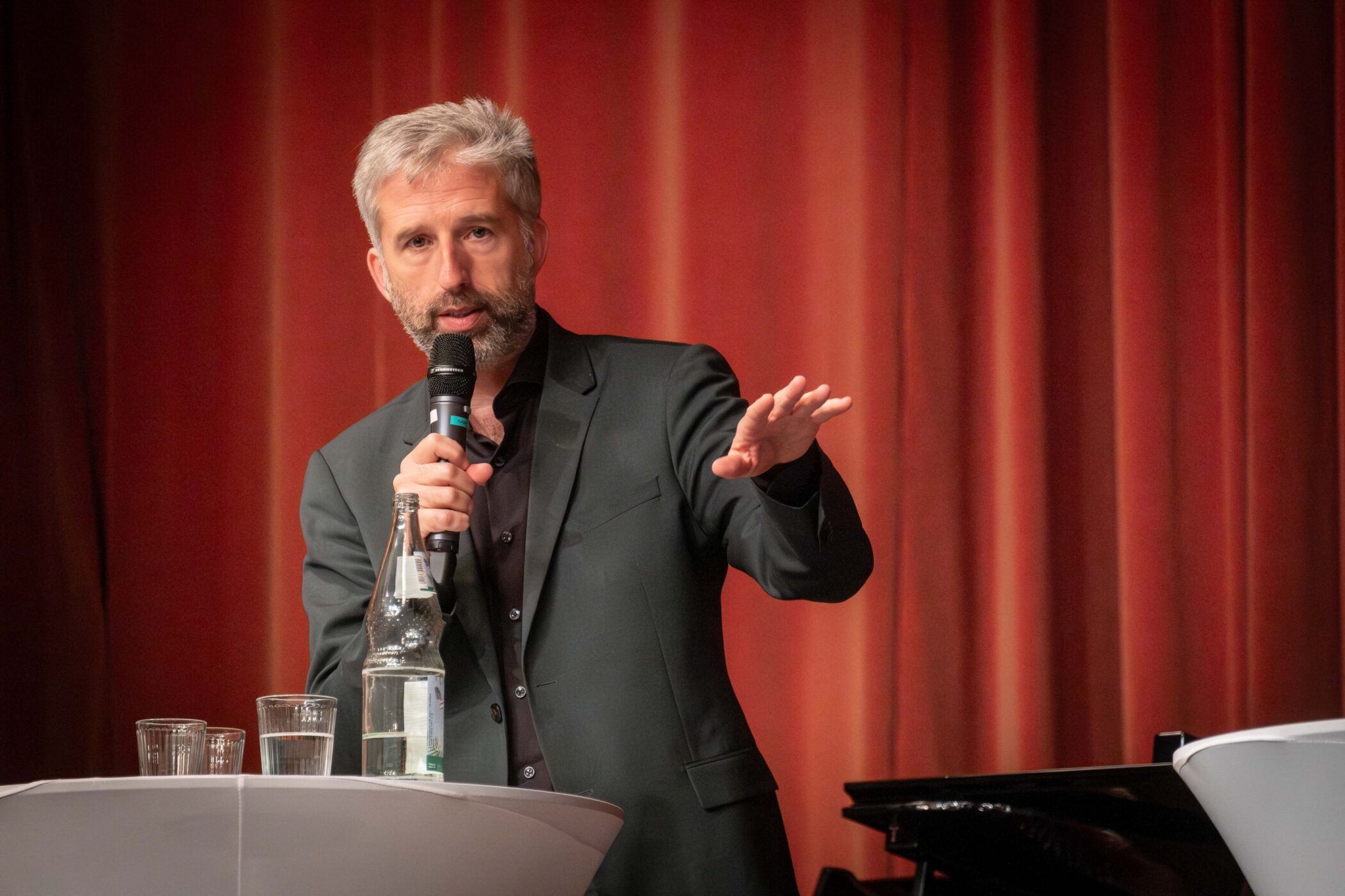 Mann mit Mikrofon spricht, steht an einem Tisch mit Flaschen und Gläsern, roter Vorhang.