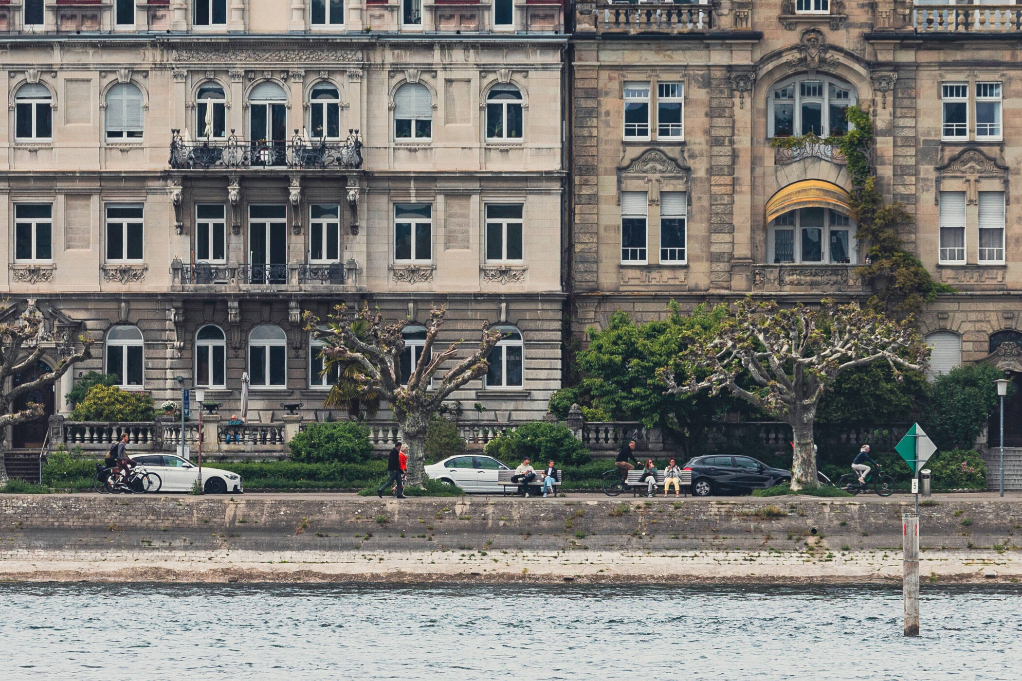 Zwei historische Gebäude mit Balkonen, davor Bäume und Menschen am Flussufer.