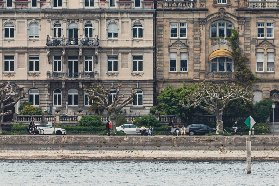 Zwei historische Gebäude mit Balkonen, davor Bäume und Menschen am Flussufer.