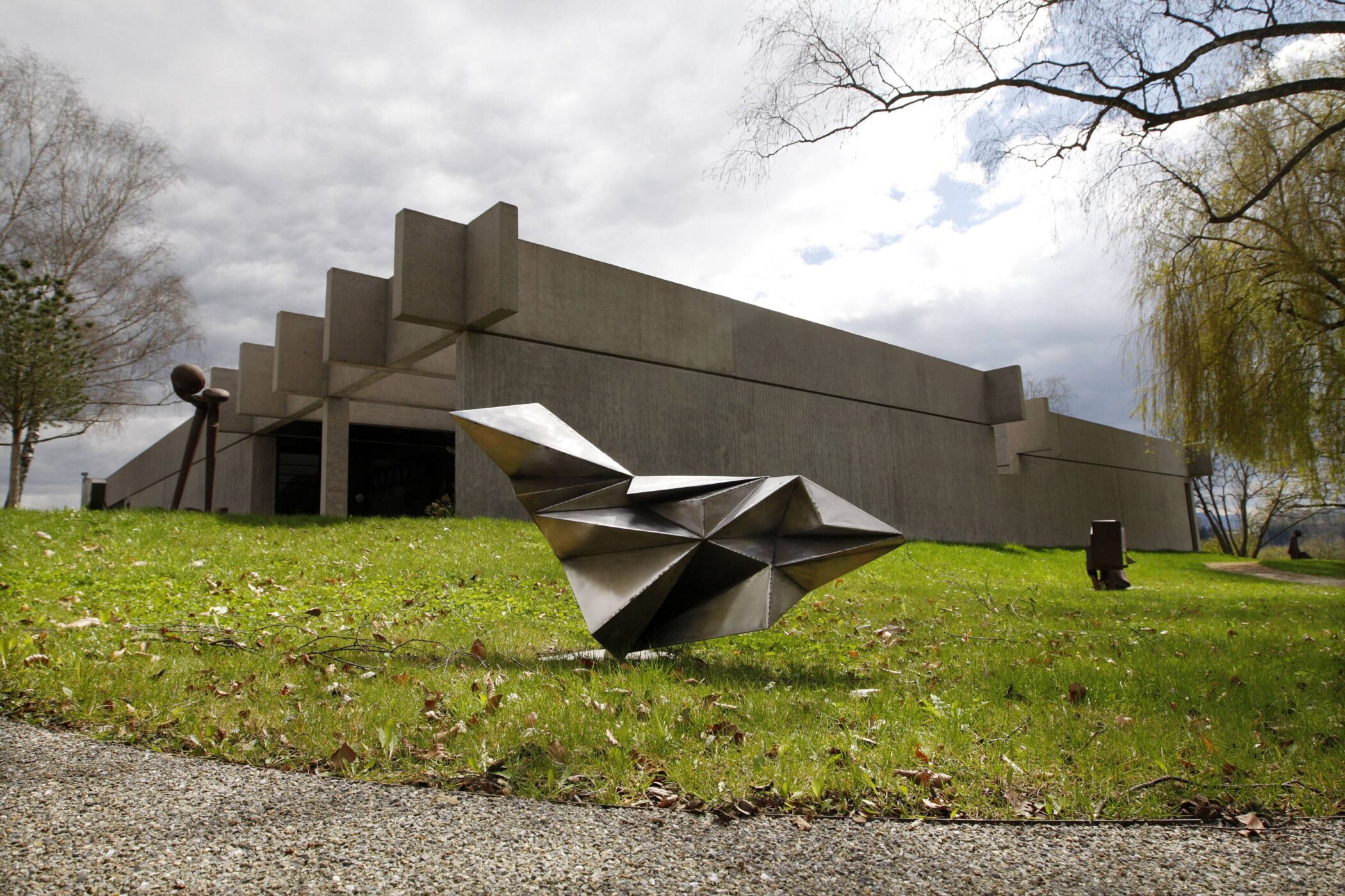 Betongebäude mit moderner Skulptur auf Rasen, Bäume im Hintergrund.