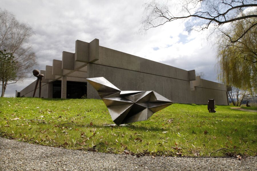Betongebäude mit moderner Skulptur auf Rasen, Bäume im Hintergrund.