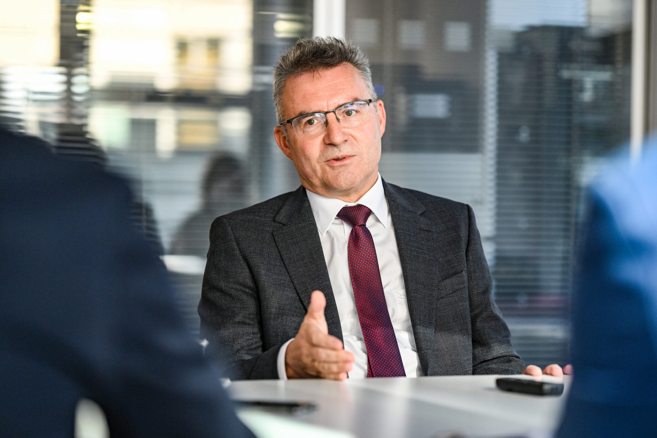 Mann im Anzug spricht gestikulierend am Tisch, unscharfe Personen im Vordergrund.