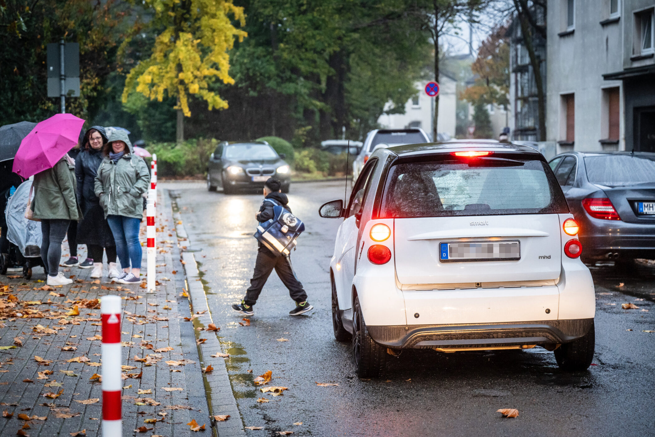 Kind überquert Straße hinter weißem Auto, vier Personen mit Regenschirmen am Gehweg.
