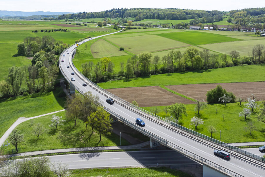 Mehrspurige Straße auf Brücke über grüne Felder, mehrere Autos unterwegs.