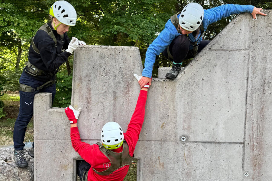 Drei Personen mit Helmen klettern an einer Betonwand, eine hilft der anderen hoch.