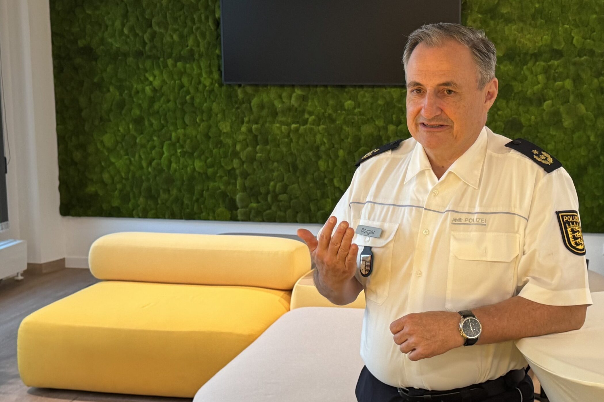 Ein Polizist in Uniform steht in einem Raum mit grüner Wand und gelben Sofas.
