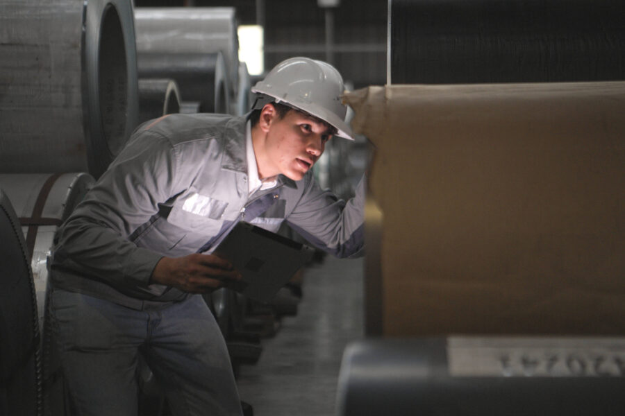 Person mit Helm inspiziert große Metallrollen in einer Fabrikhalle.