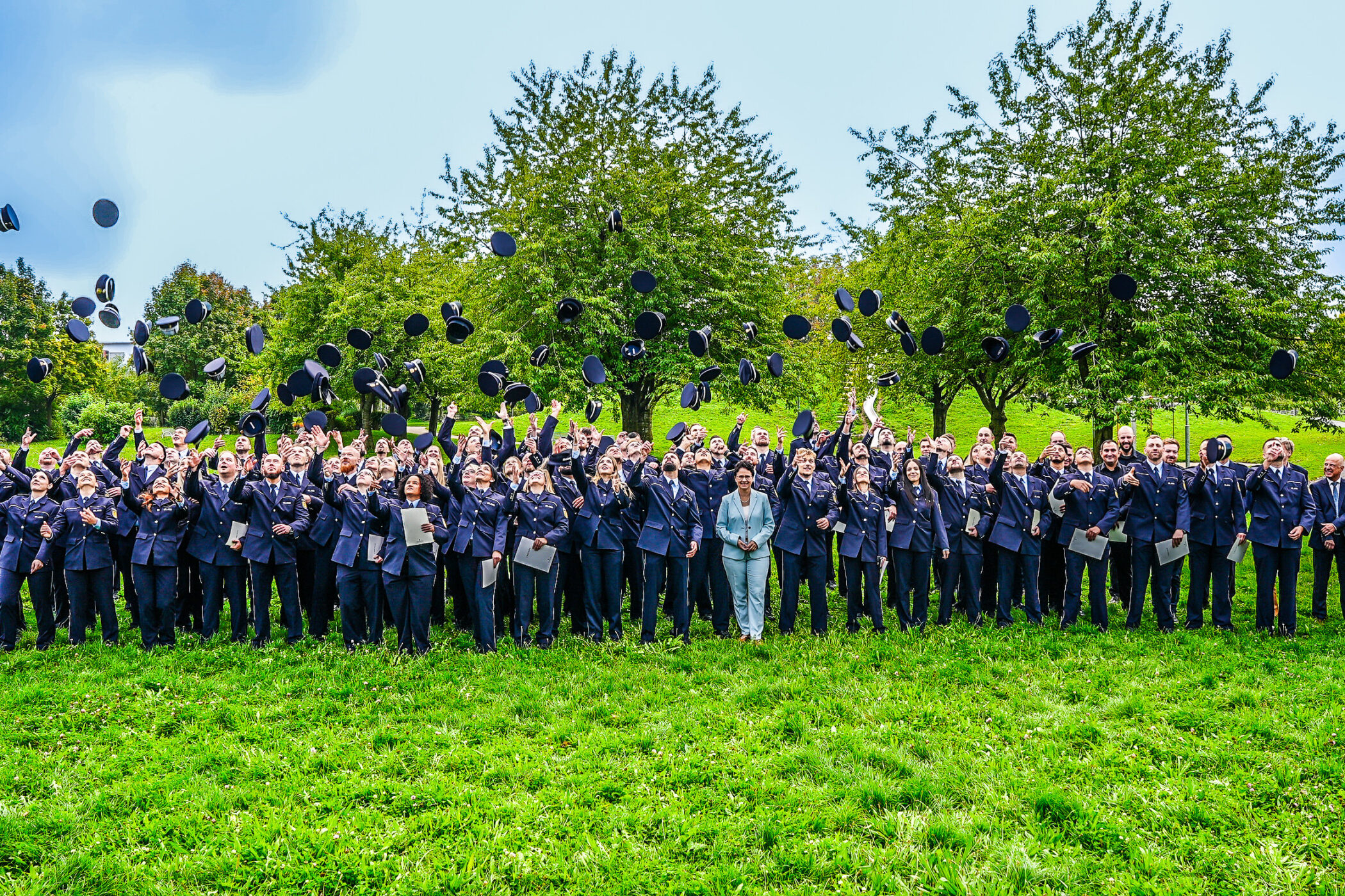 Gruppe von Menschen in Uniformen wirft Hüte in die Luft, im Freien auf einer Wiese.