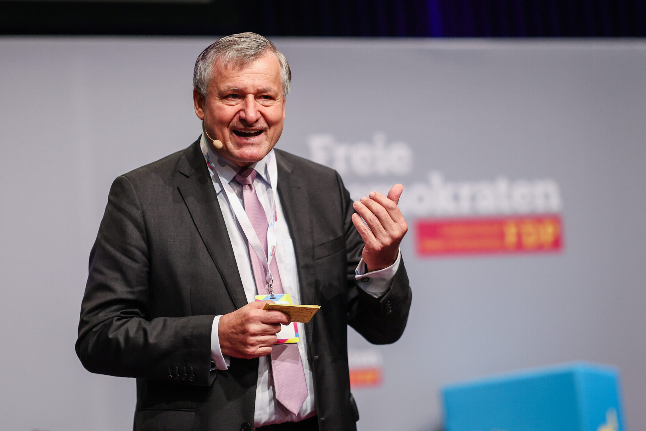 Älterer Mann im Anzug spricht auf einer Bühne, Mikrofon am Ohr.