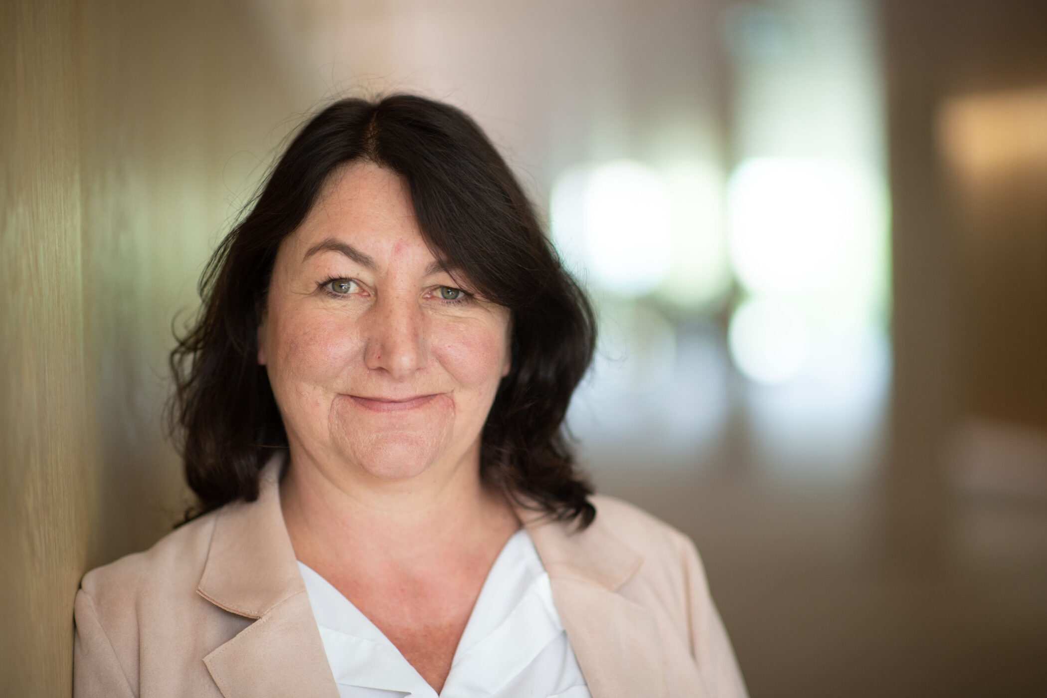 Frau mit dunklem Haar und beigem Blazer lächelt in die Kamera.