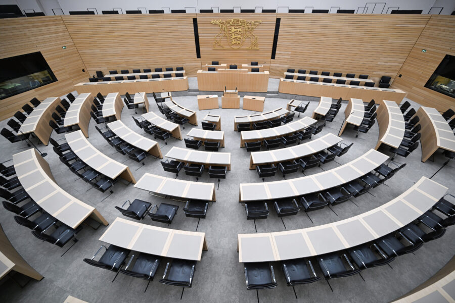 Leerer Sitzungssaal mit halbkreisförmigen Tischen und schwarzen Stühlen.