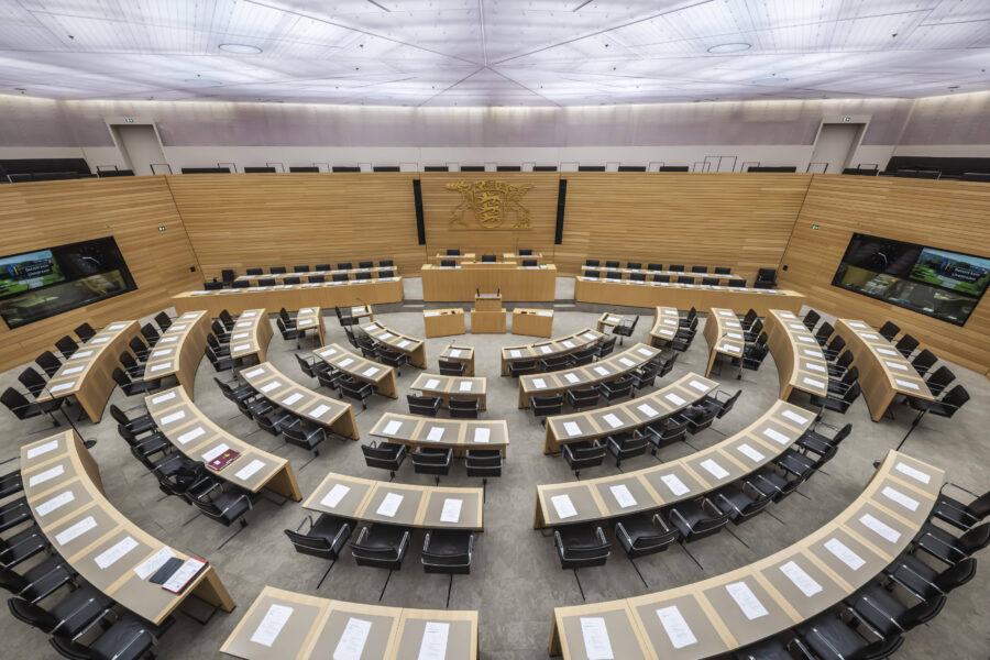 Leerer Sitzungssaal mit halbkreisförmigen Tischen und schwarzen Stühlen.