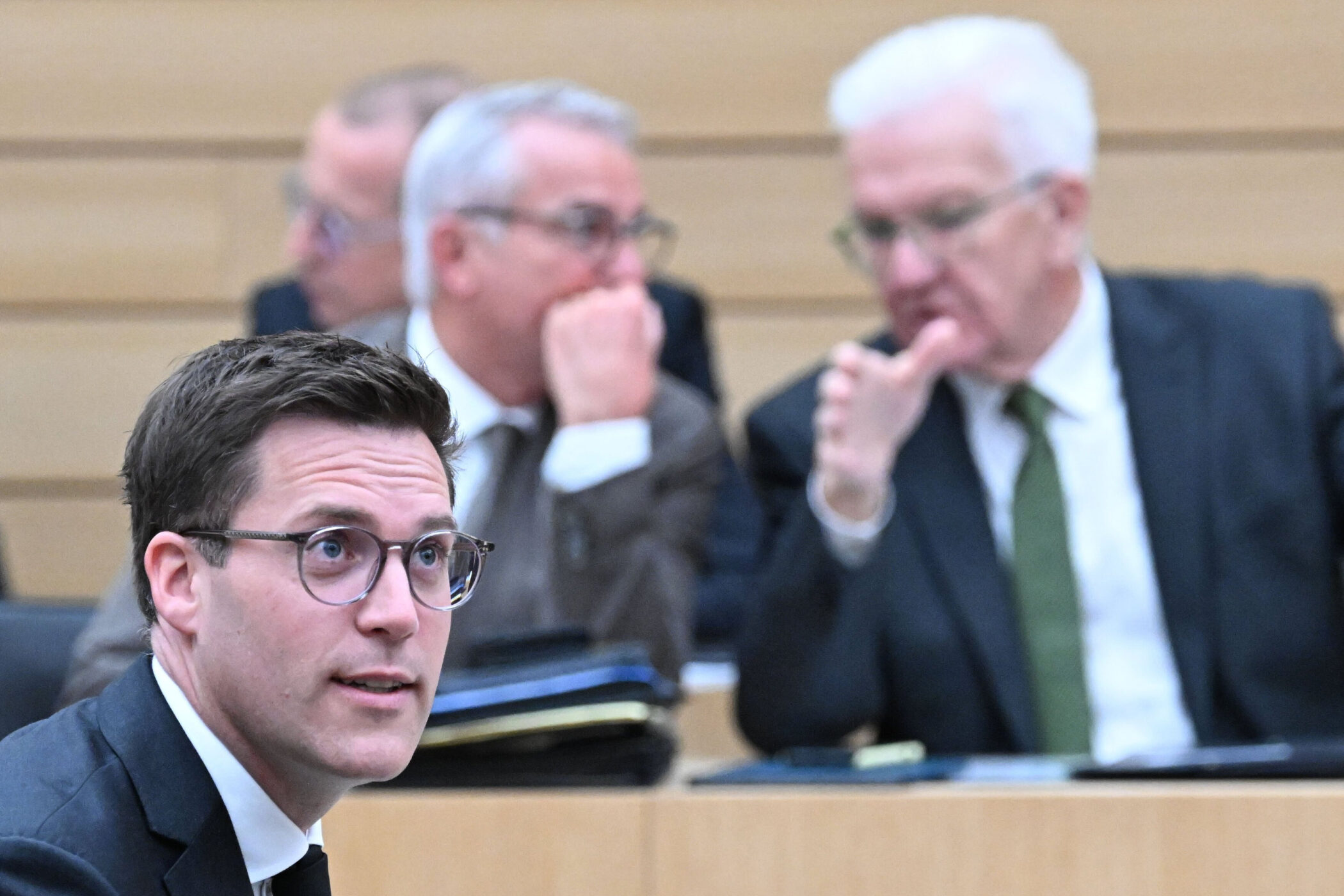 Drei Männer in Anzügen sitzen in einem Raum, einer spricht gestikulierend.