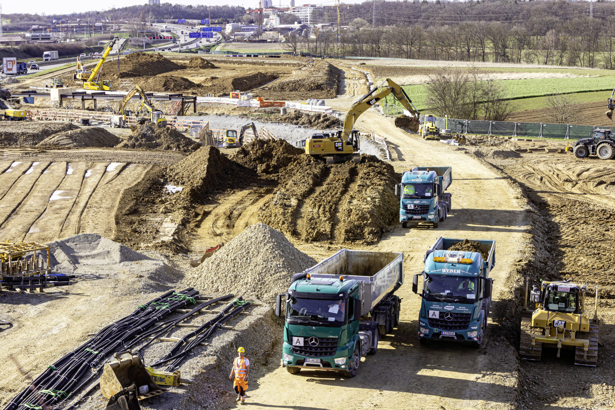 Baustelle mit Baggern, Lastwagen und Bauarbeiter in orangefarbener Schutzkleidung.