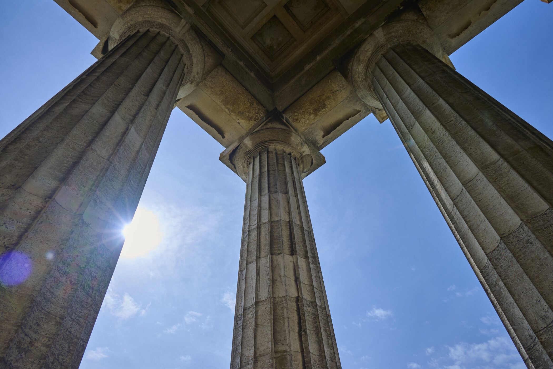 Drei steinerne Säulen von unten, blauer Himmel und Sonne im Hintergrund.
