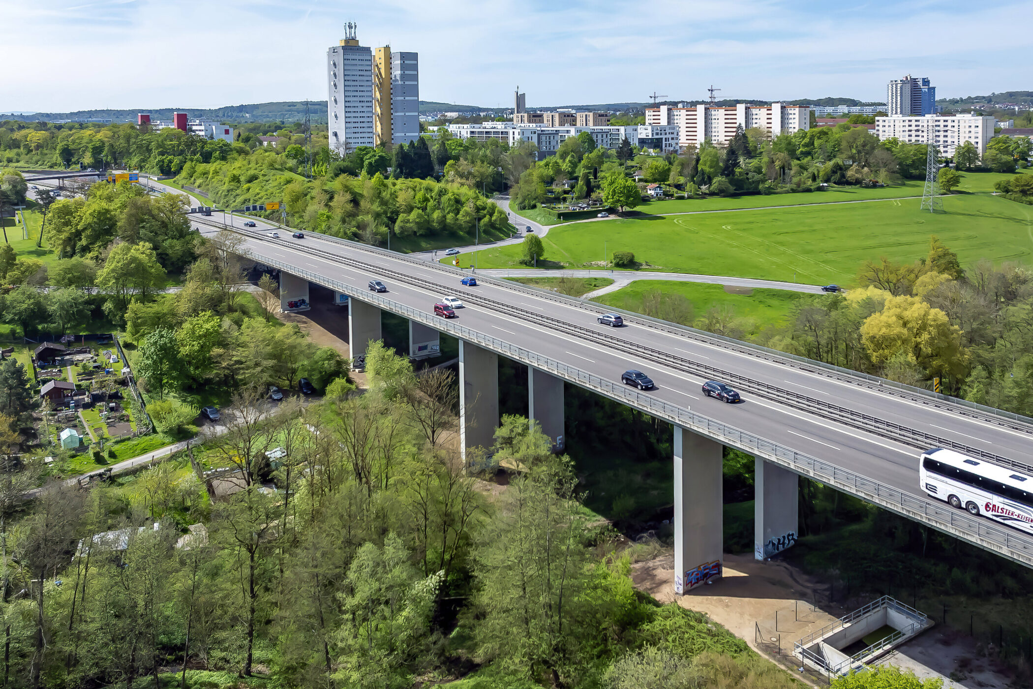 Autobahnbrücke mit Autos und Bus, umgeben von Bäumen und Wohngebäuden im Hintergrund.