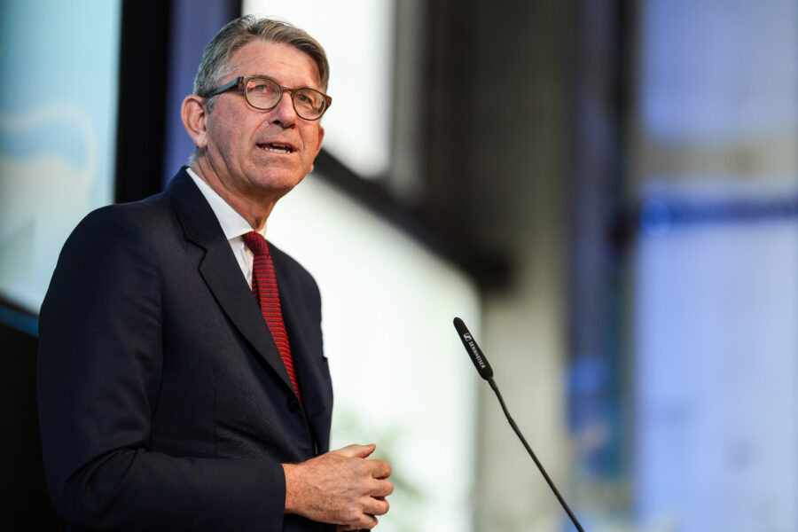 Älterer Mann mit Brille spricht an einem Mikrofon, trägt Anzug und rote Krawatte.