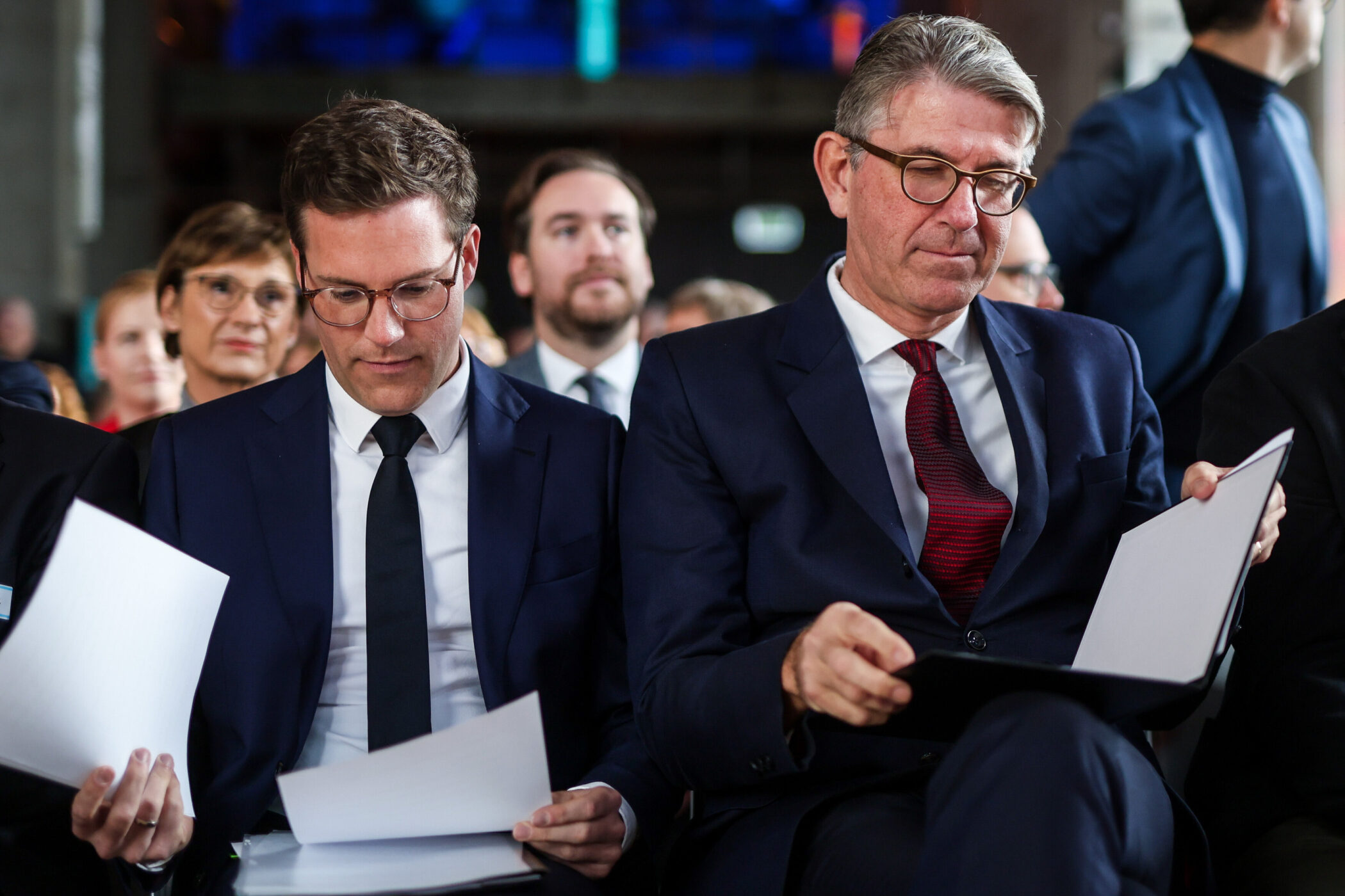 Zwei Männer in Anzügen sitzen nebeneinander und lesen Dokumente.