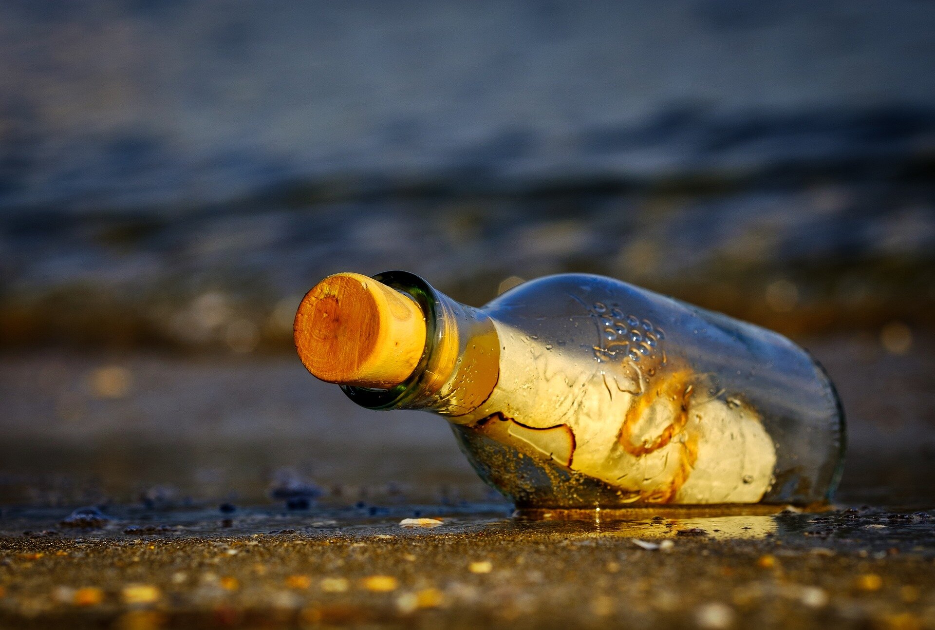 Flasche mit Korken und Papier am Strand, Wasser im Hintergrund.