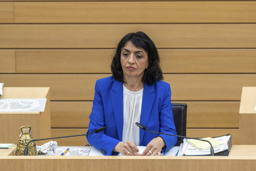 Eine Frau in blauem Blazer sitzt an einem Tisch mit Mikrofonen und Papieren.