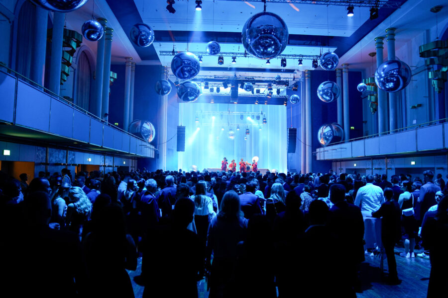 Menschenmenge in einem Saal mit Bühne und Spiegelkugeln an der Decke.